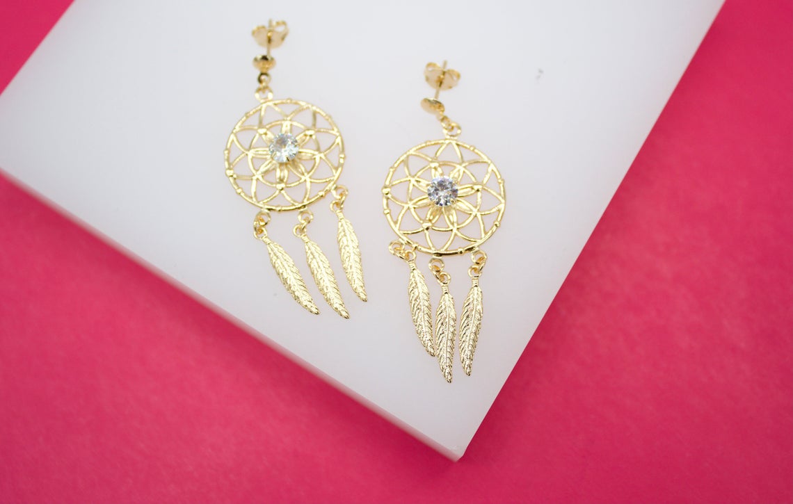 A pair of gold dreamcatcher earrings with dangling feathers and central zirconia stones rests on a white display against a vibrant pink background.