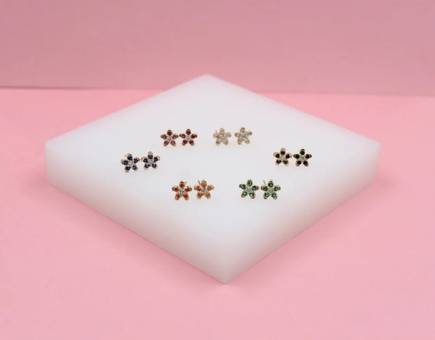 A collection of colorful flower-shaped stud earrings featuring gemstone petals rests on a white display block against a pink background.