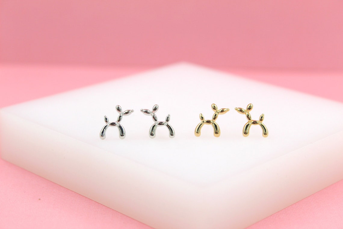 Four shiny balloon dog earrings in silver and gold rest on a white display against a soft pink backdrop.