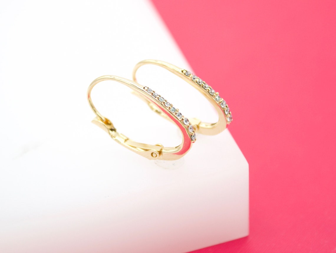 A pair of delicate gold hoop earrings with sparkling cubic zirconia stones rests on a white and pink display stand.