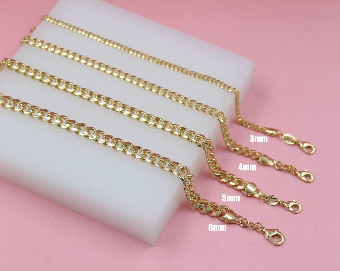 Four gold-plated cuban link chains in varying widths of 3mm, 4mm, 5mm, and 6mm rest on a white display block against a pink background.