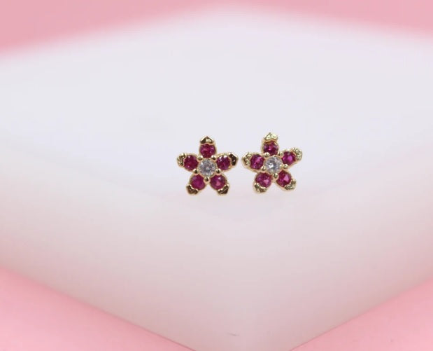A pair of gold flower stud earrings features sparkling ruby petals and a clear center crystal resting on a soft white display surface.