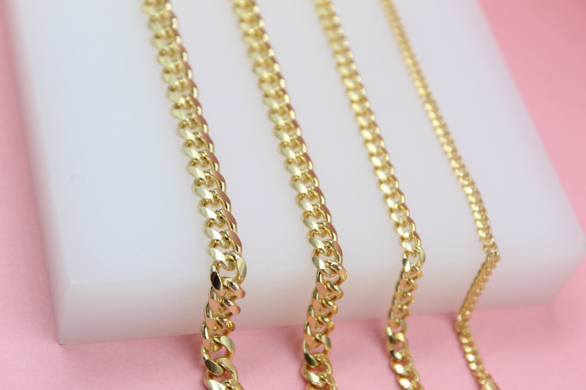 Four different thicknesses of gold cuban link chains rest on a white display against a pink background.