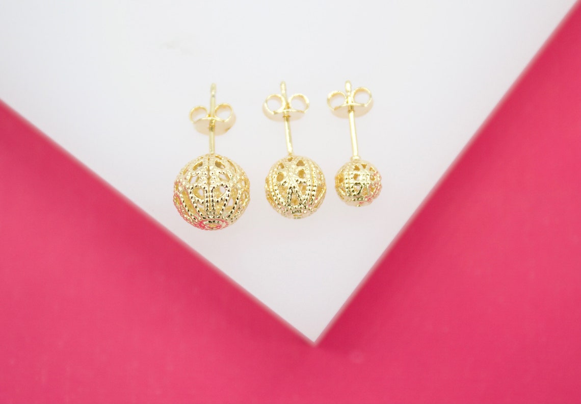Three gold filigree ball stud earrings of descending sizes rest on a white display stand against a vibrant pink background.