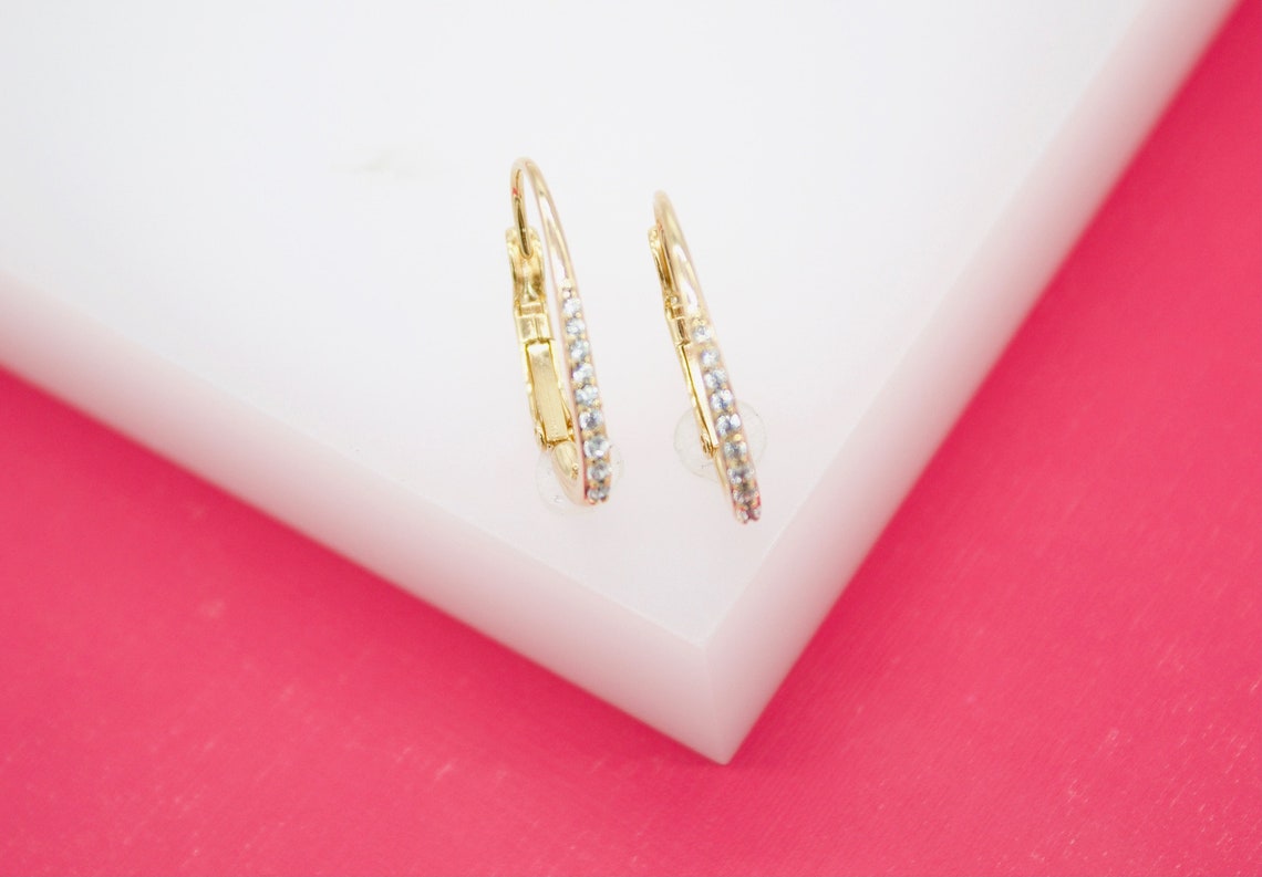 A pair of delicate gold huggie hoop earrings featuring sparkling pave-set diamonds rests on a white display against a vibrant pink background.