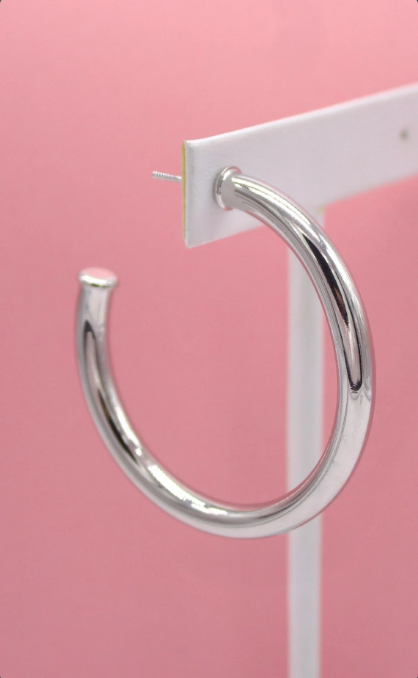 A polished silver hoop earring hangs on a white display card against a soft pink background.