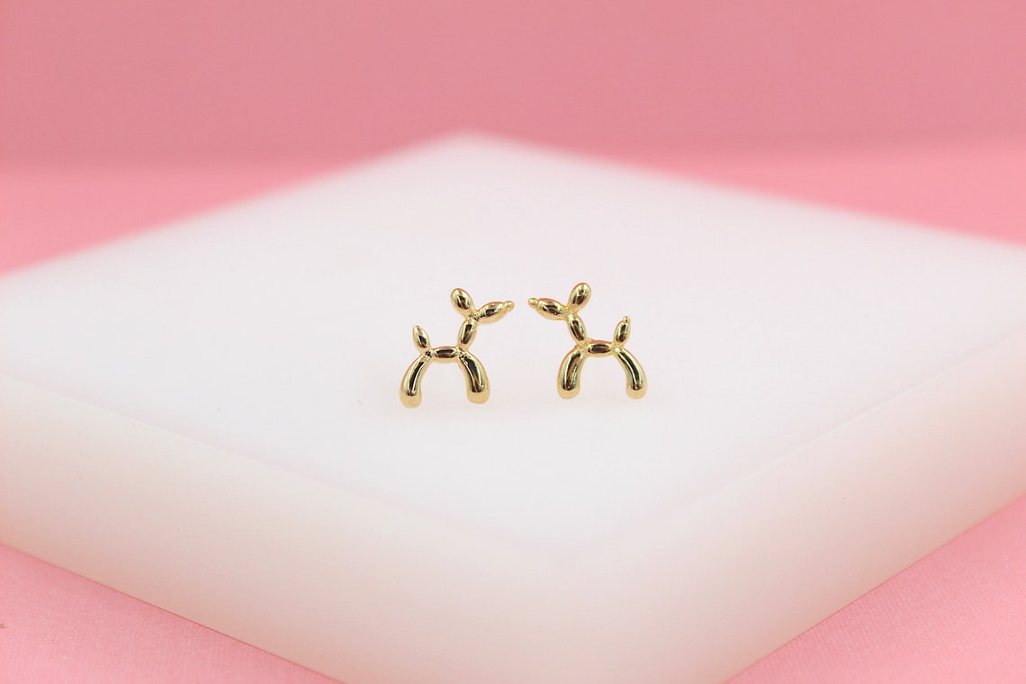 A pair of cute gold balloon dog stud earrings rests on a white display surface against a pink background.