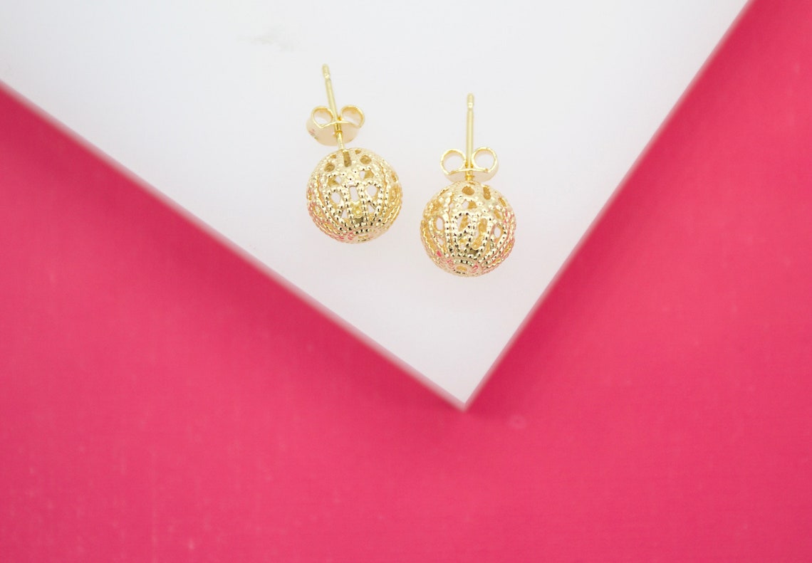 A pair of intricate gold filigree ball stud earrings rests on a white display against a vibrant pink background.