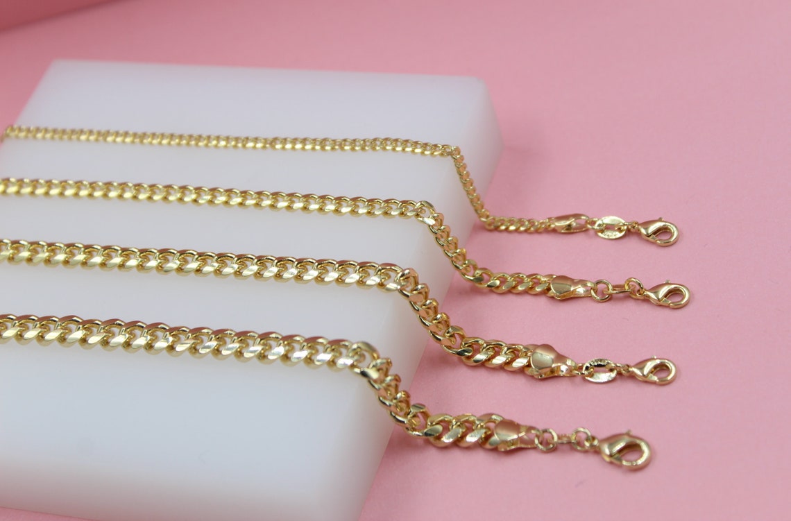 Four different-sized gold cuban link necklaces display their clasps on a white display block against a pink background.