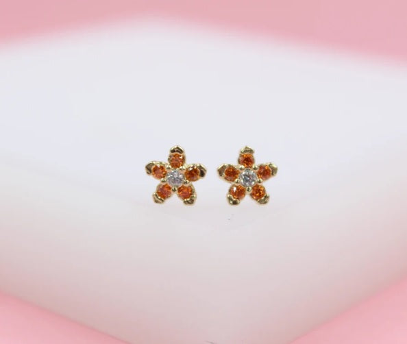 A pair of gold flower-shaped stud earrings features orange crystal petals and a clear center stone against a soft white and pink background.