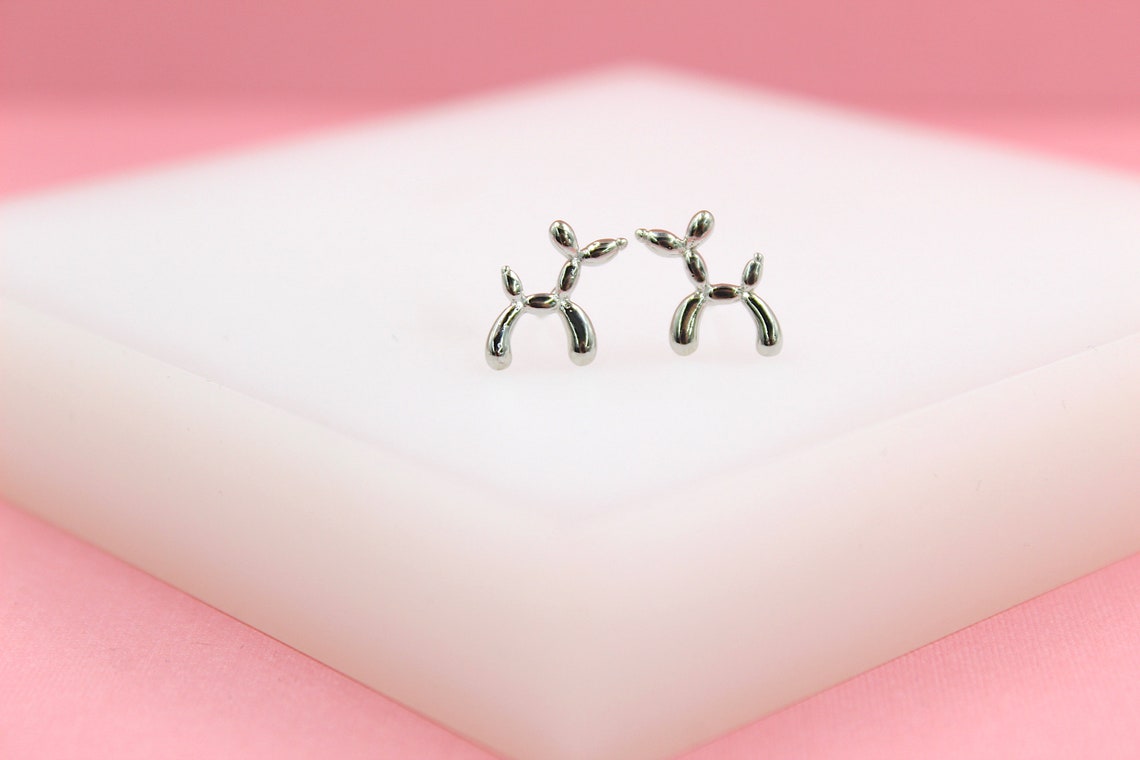 A pair of shiny silver balloon dog stud earrings rests on a white display against a pink background.