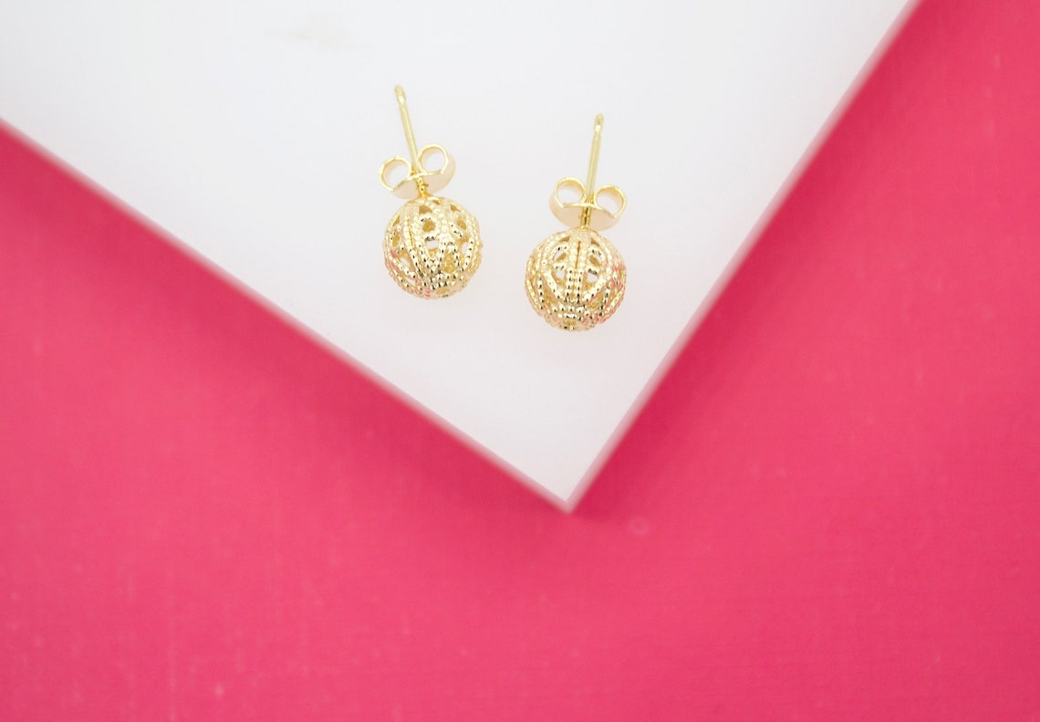 A pair of intricate gold filigree ball stud earrings rests on a white display against a vibrant pink background.