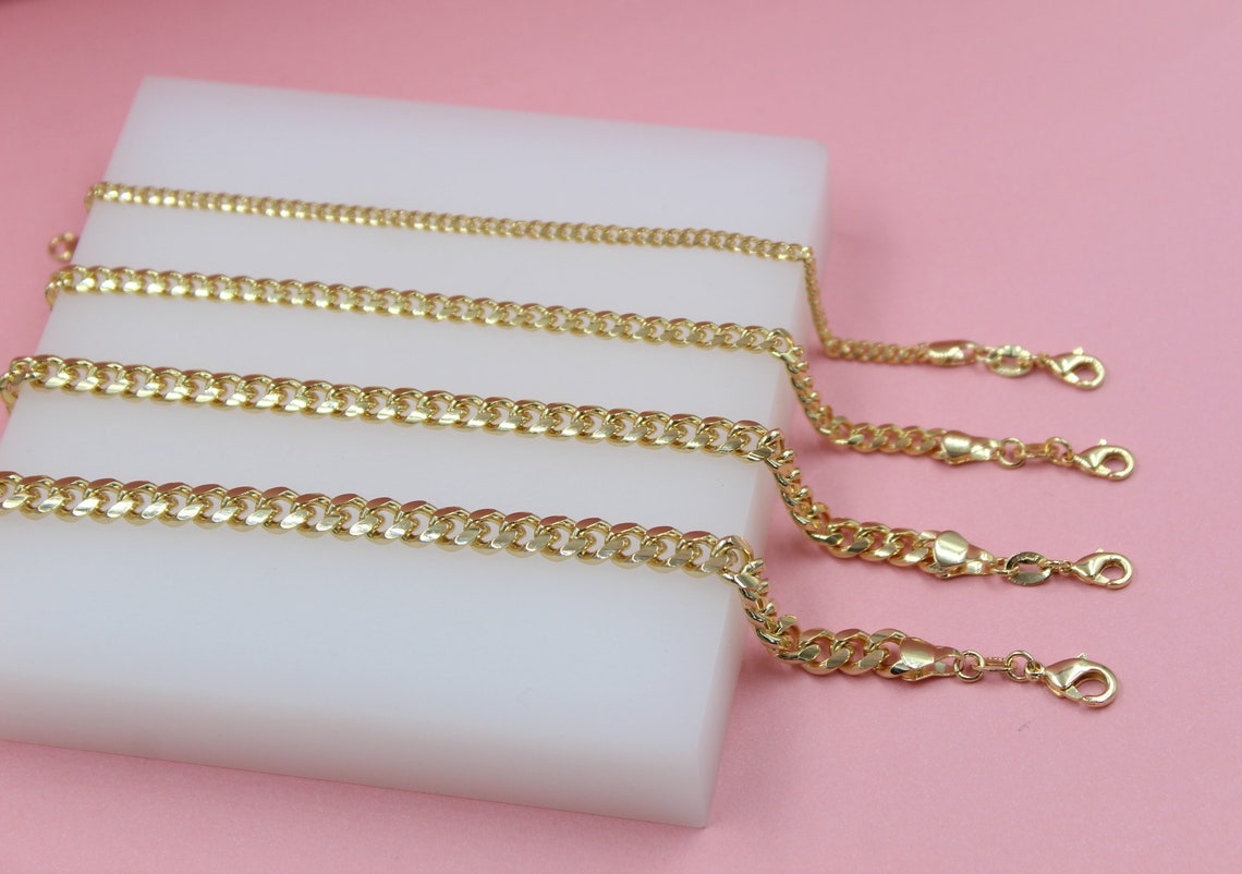 Four gold cuban link chains in varying widths rest neatly on a white display block against a soft pink background.