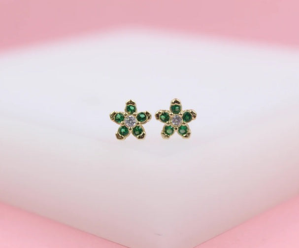 A pair of gold flower stud earrings featuring green crystal petals and a sparkling white center stone rests on a soft white surface.