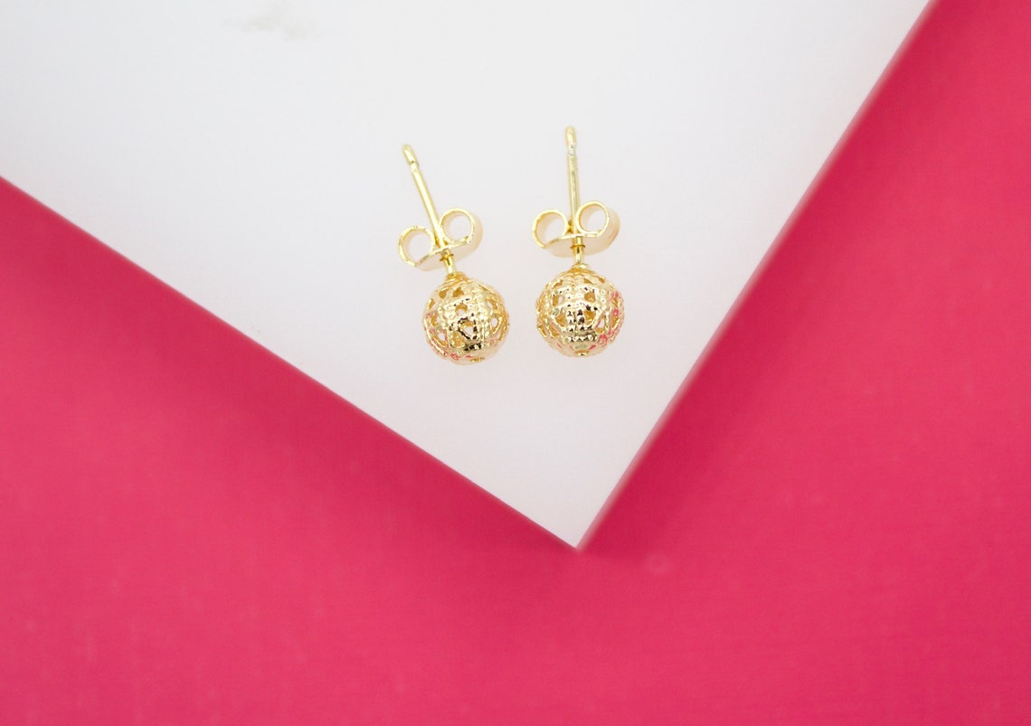 A pair of gold filigree ball stud earrings rests on a white and bright pink background.