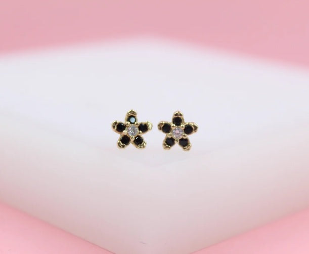 Two gold flower stud earrings feature dark petals and a clear center stone against a soft pink background.