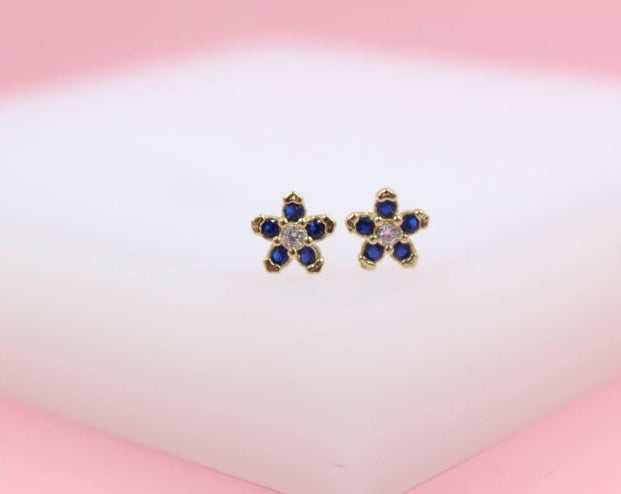 A pair of gold flower stud earrings features dark blue cubic zirconia petals and a clear center stone against a soft white background.