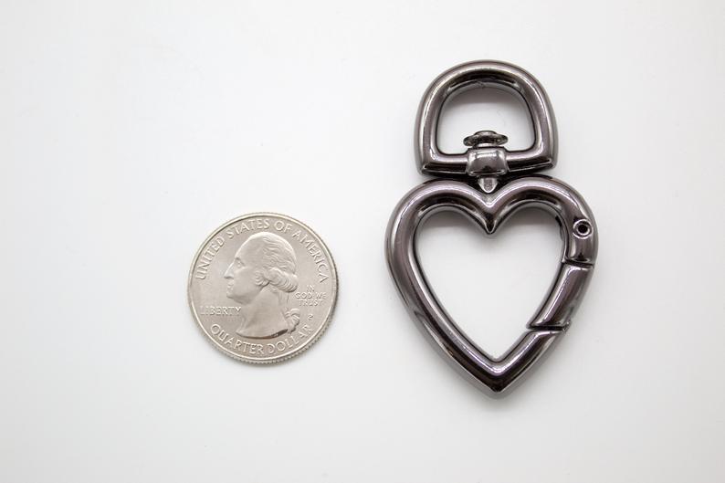 A gunmetal heart-shaped swivel lobster clasp lies next to a US quarter coin on a white surface to show its scale.