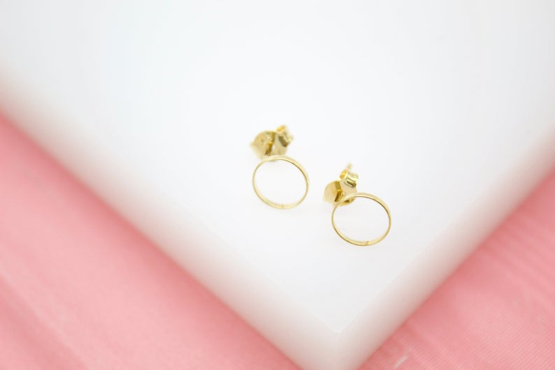 A pair of delicate gold circle stud earrings rests on a white and pink surface.