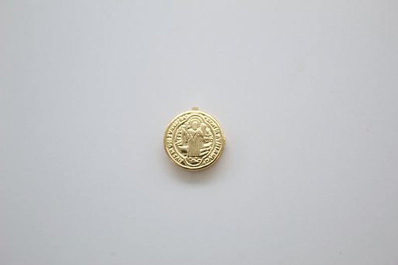 A small round gold Saint Benedict medal lies centered on a plain white background.