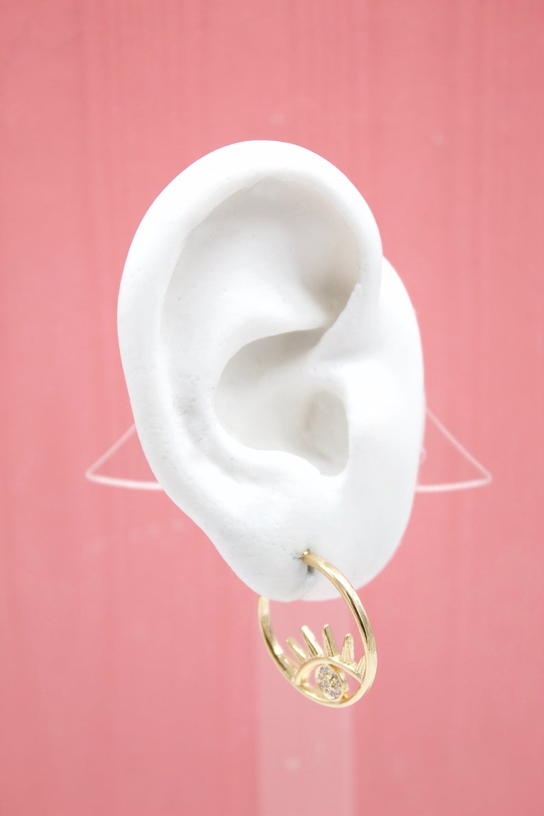 A white mannequin ear displays a gold evil eye hoop earring against a pink background.