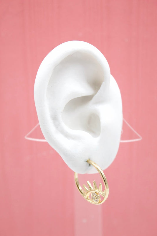 A white mannequin ear displays a gold evil eye hoop earring against a pink background.