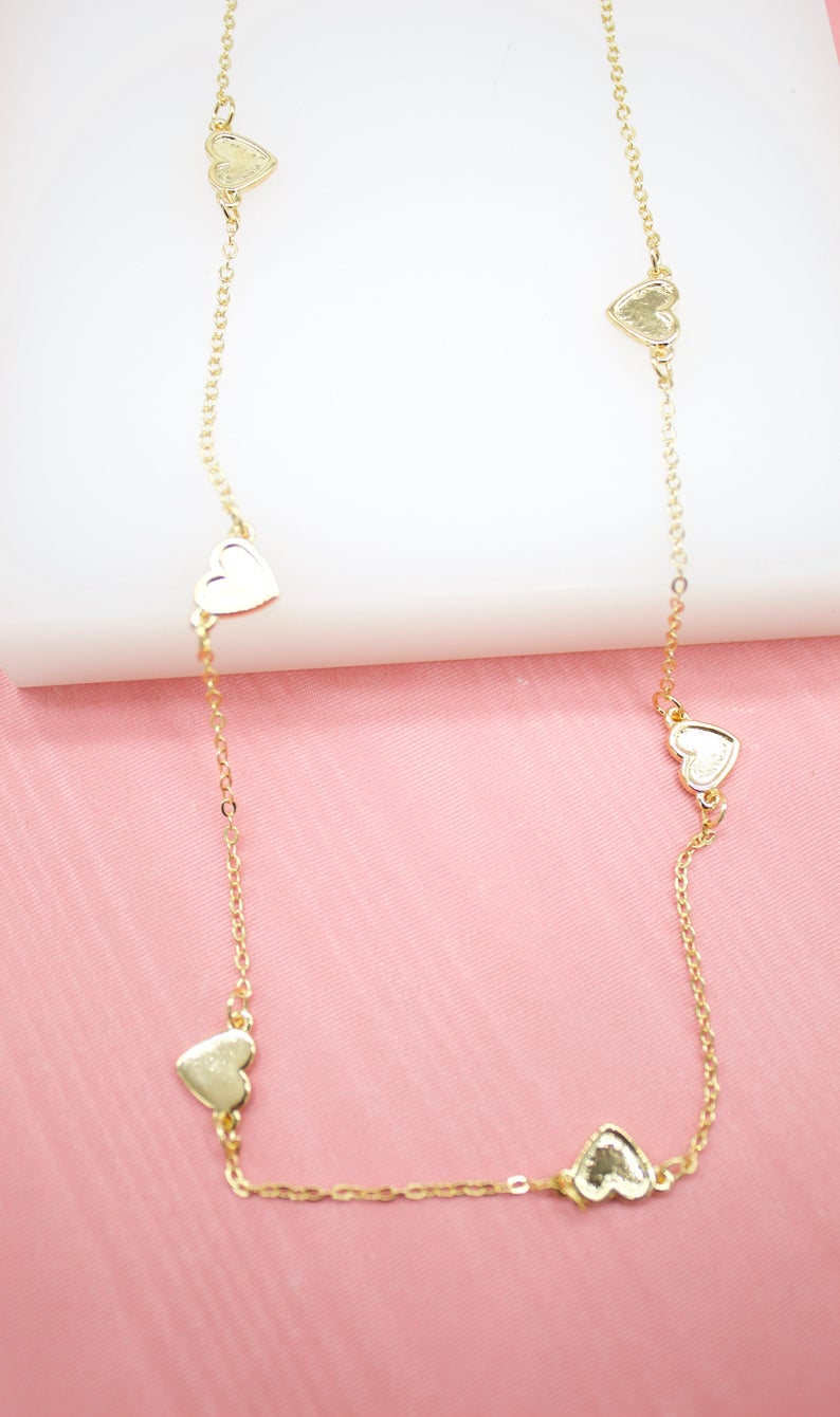 A delicate gold chain necklace featuring multiple evenly spaced heart charms drapes against a textured pink and white surface.
