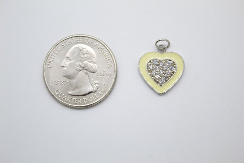 A light yellow and silver heart charm rests next to a US quarter coin for size comparison.
