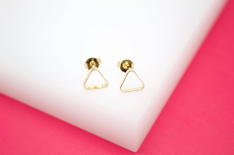A pair of minimalist gold triangle stud earrings rests on a white display surface against a vibrant pink background.