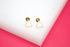 A pair of minimalist gold triangle stud earrings rests on a white display surface against a vibrant pink background.