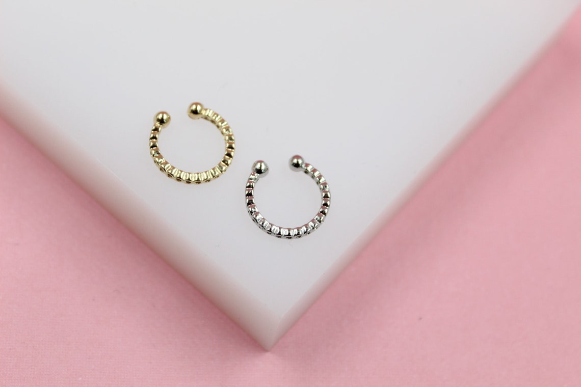 A gold ear cuff and a silver ear cuff rest side by side on a white surface with a pink background.