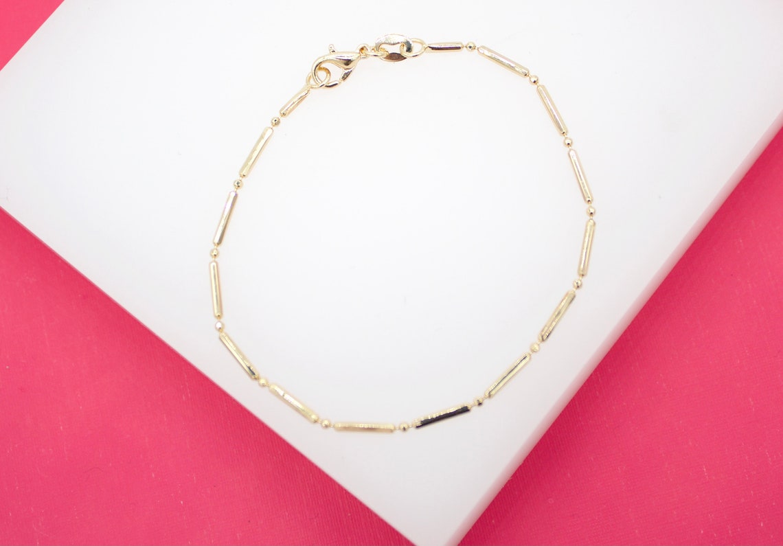 A delicate gold link bracelet rests on a white display surface against a vibrant pink background.