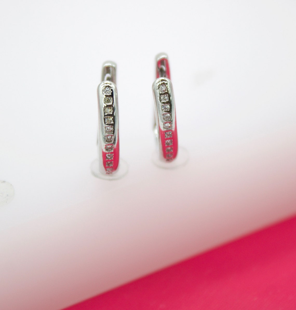 A pair of silver hoop earrings features vibrant pink enamel and a row of sparkling diamond accents displayed on a white surface.