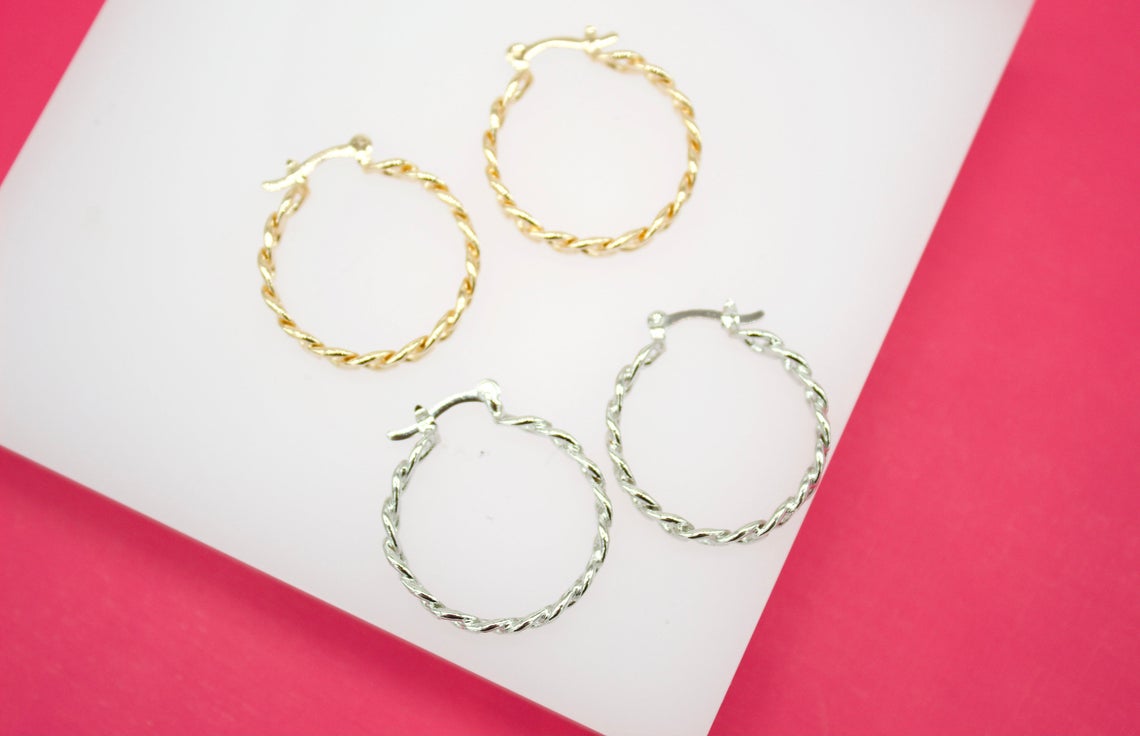 Two gold and two silver twisted hoop earrings rest on a white display surface against a bright pink background.