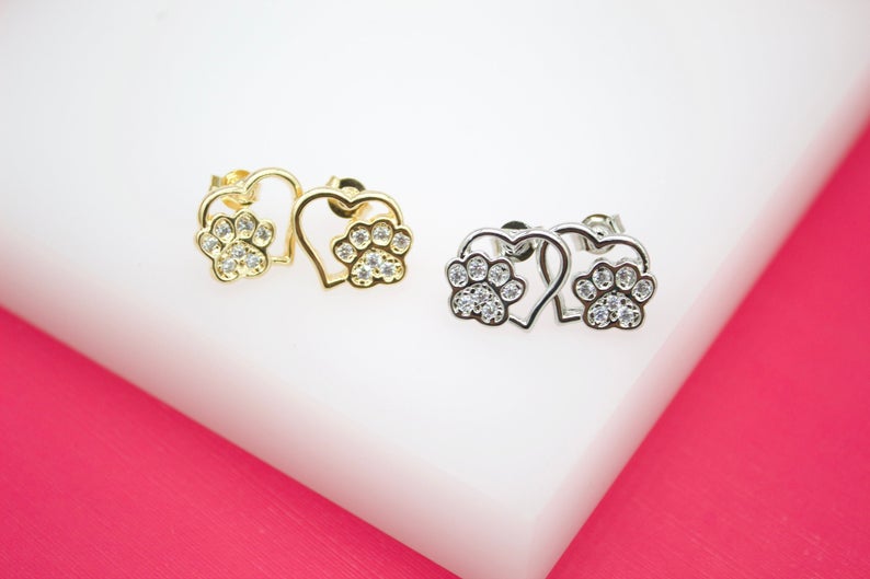 A pair of gold and silver heart-shaped stud earrings featuring crystal-embellished dog paw prints rests on a white and pink display surface.