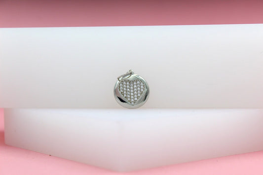 A silver circular pendant featuring a pavé-set crystal heart rests on a white display block against a soft pink background.