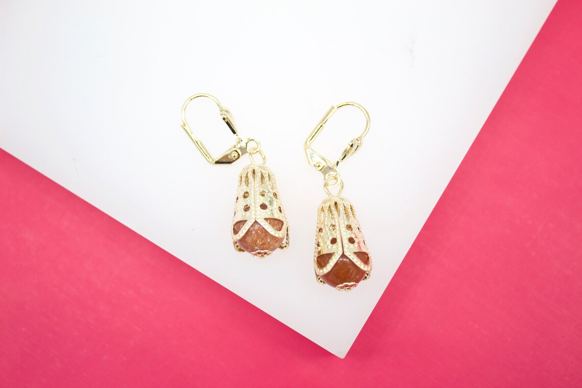 A pair of gold leverback earrings features intricate filigree cages cradling shimmering reddish-brown gemstones.
