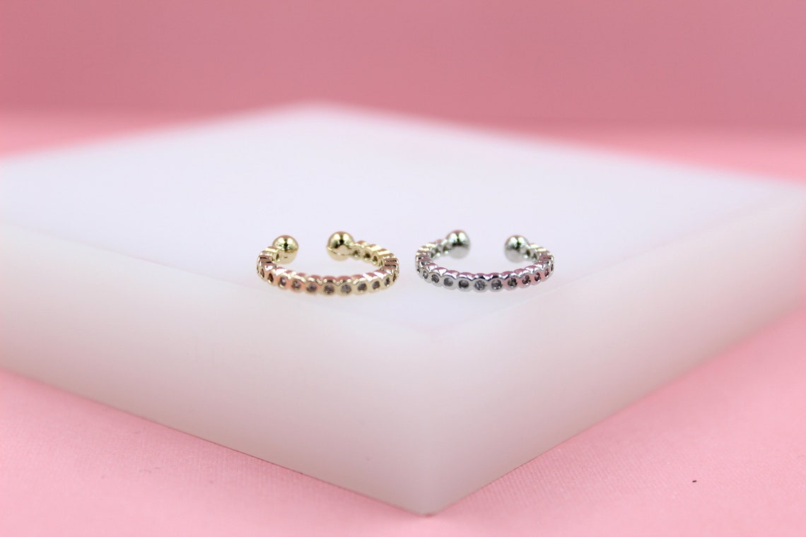 A gold and a silver ear cuff with sparkling zirconias rest side by side on a white display block against a soft pink background.