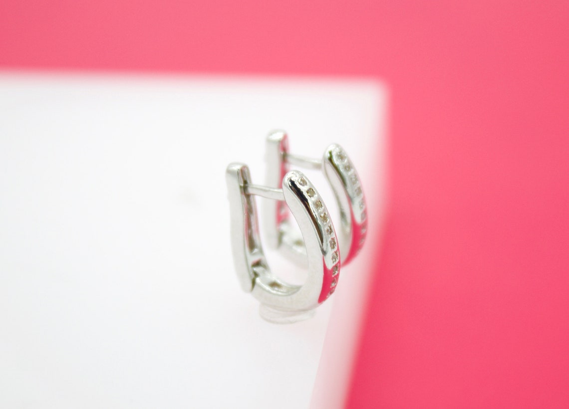 A pair of silver hoop earrings encrusted with small sparkling diamonds sits upright on a white platform against a vibrant pink background.