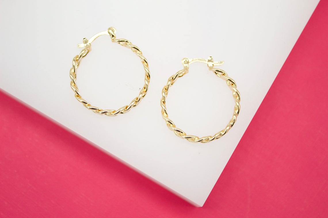 A pair of polished gold twist hoop earrings rests on a white display surface against a vibrant pink background.