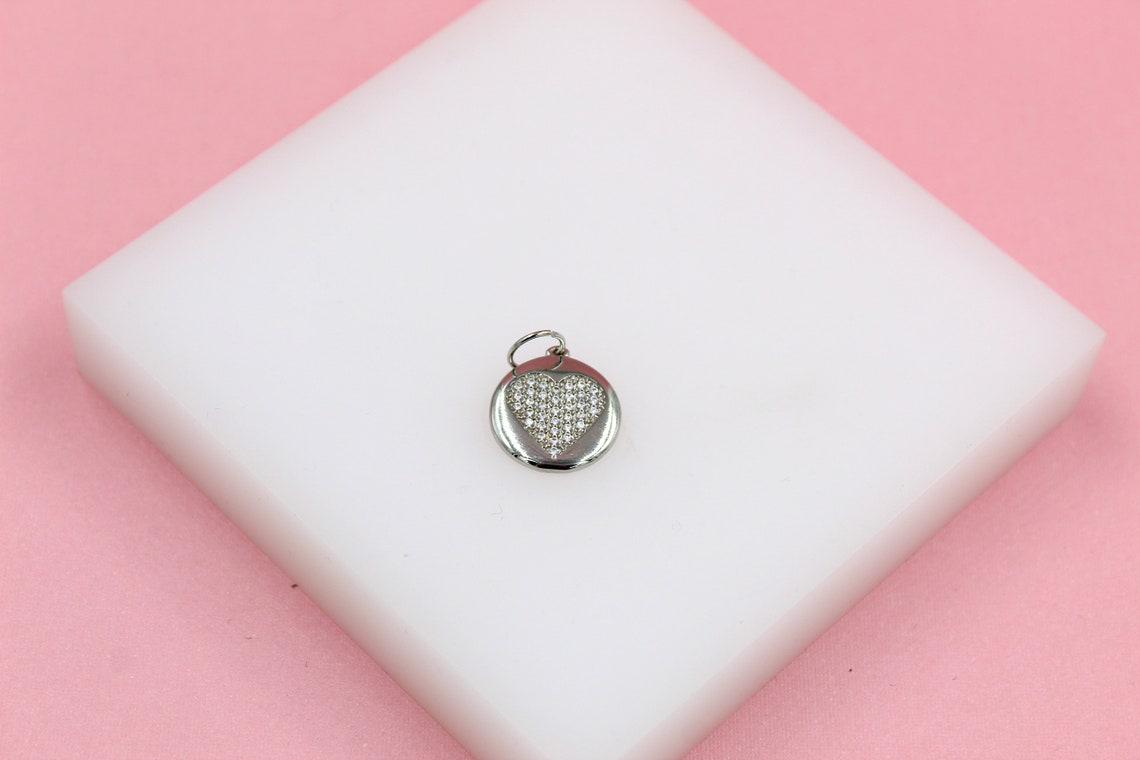 A silver pendant featuring a pavé-set diamond heart rests on a white display block against a soft pink background.