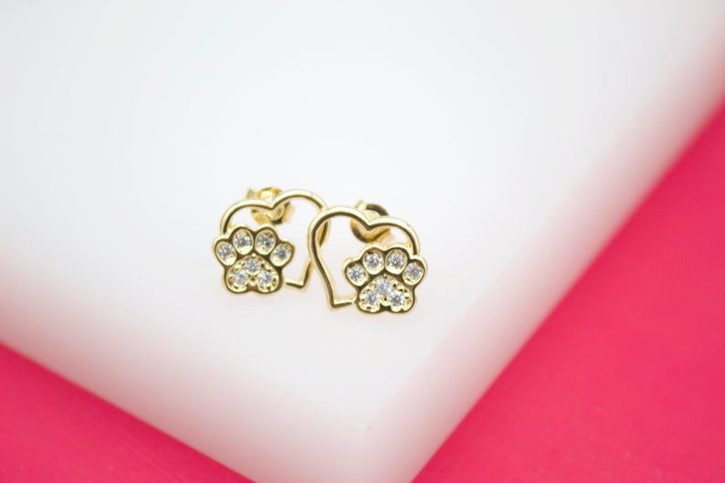 A pair of gold-plated paw print stud earrings featuring sparkling cubic zirconia stones nestled inside delicate heart frames rests on a white display surface.
