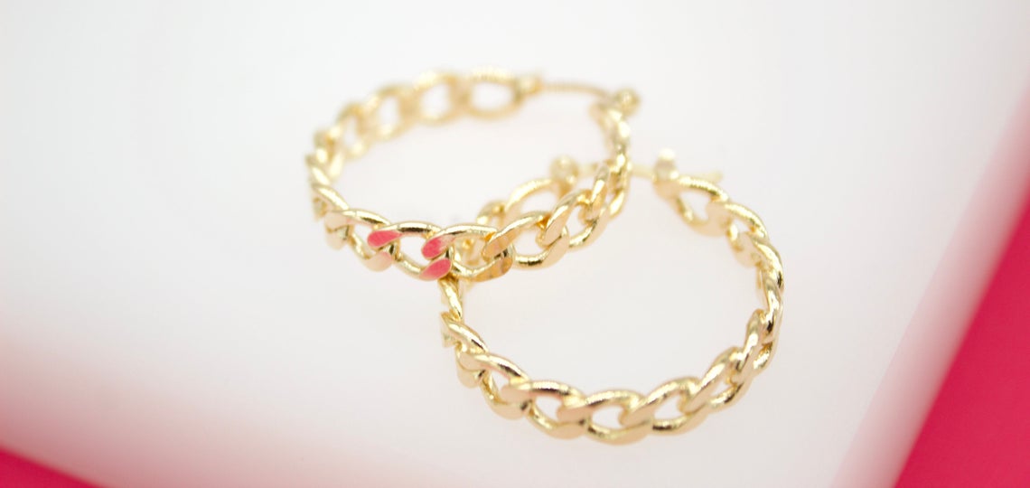Two gold chain-link hoop earrings featuring a vibrant pink accent rest against a neutral background.