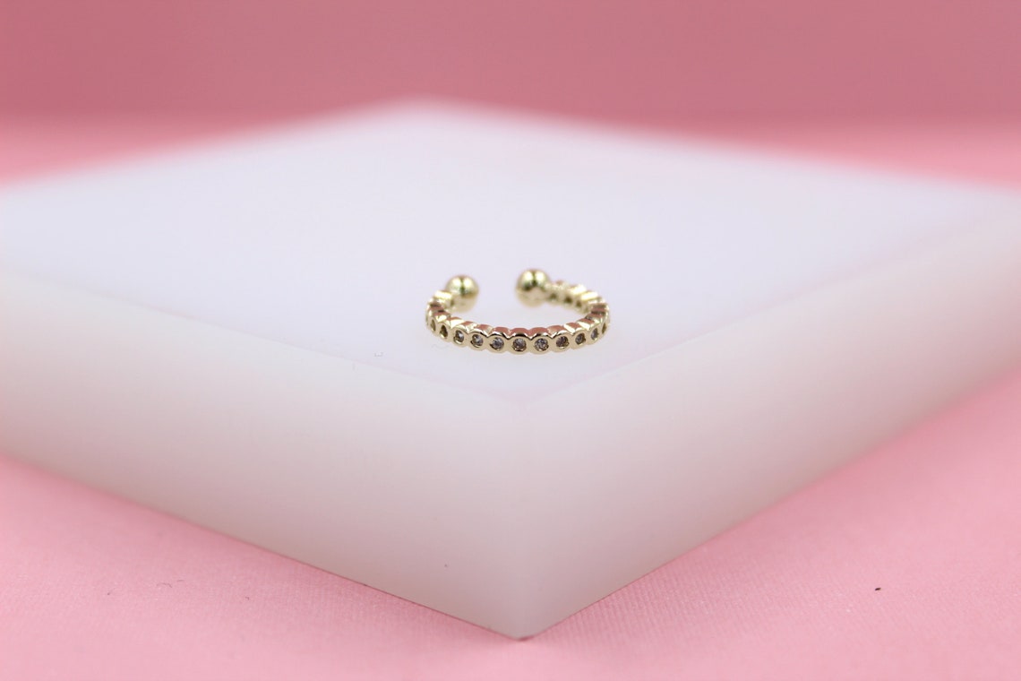 A sparkling gold ear cuff rests on a white display block against a soft pink background.