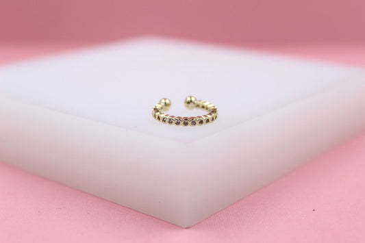 A sparkling gold ear cuff rests on a white display block against a soft pink background.