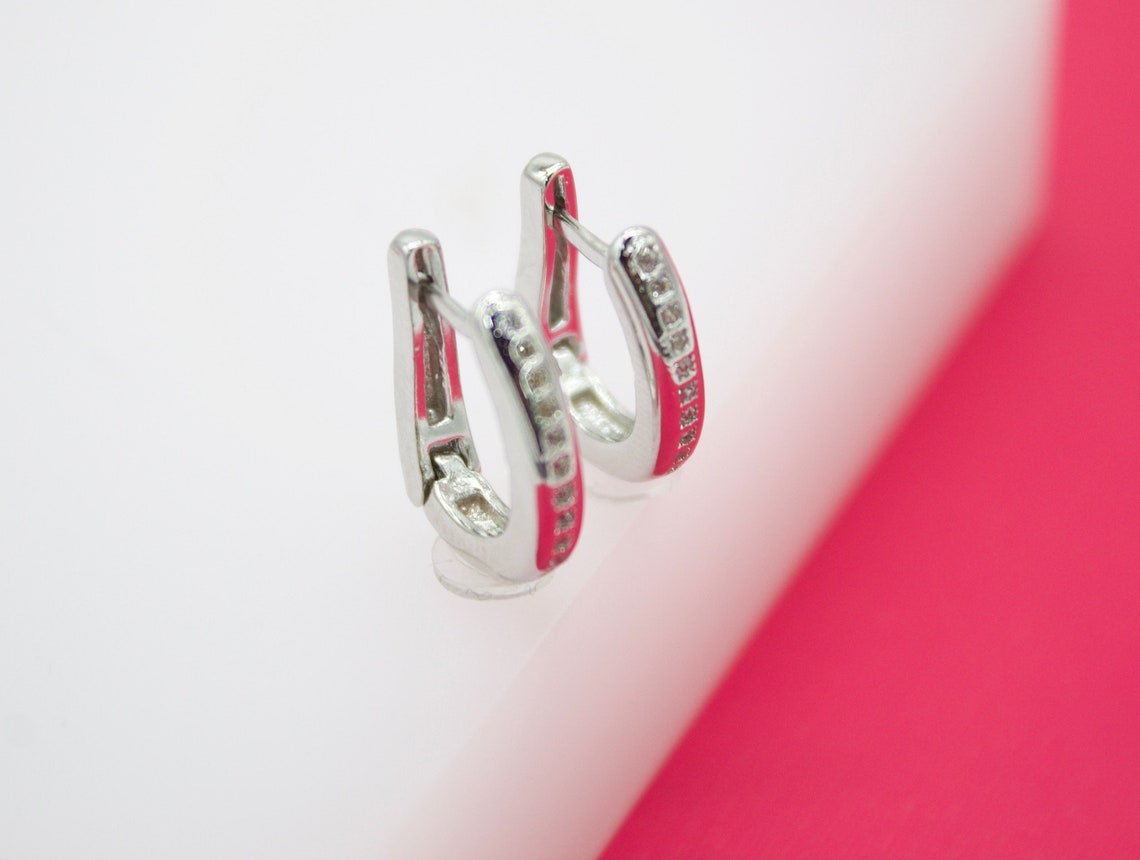 A pair of silver hoop earrings, accented with rows of sparkling pave stones and vibrant pink enamel, rests upright on a white display surface.
