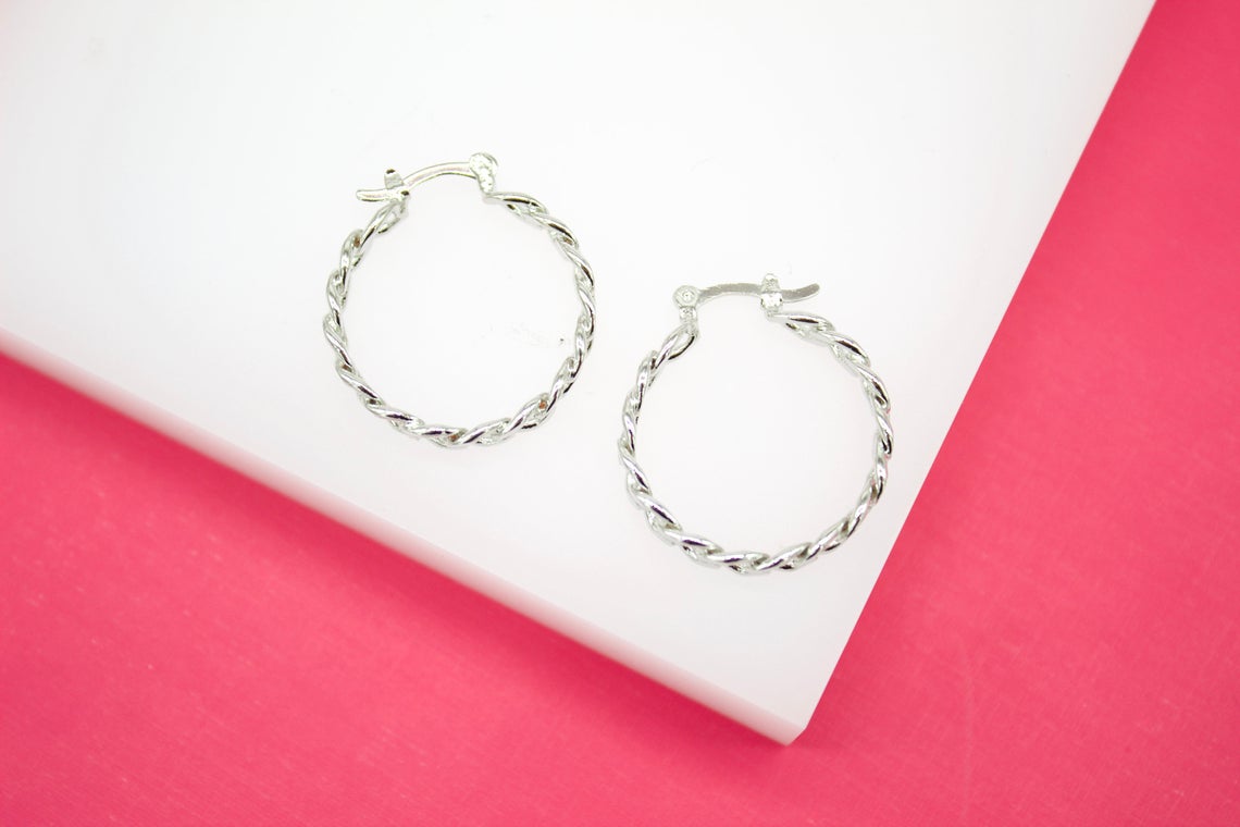 A pair of silver twisted hoop earrings rests on a white display block against a vibrant pink background.