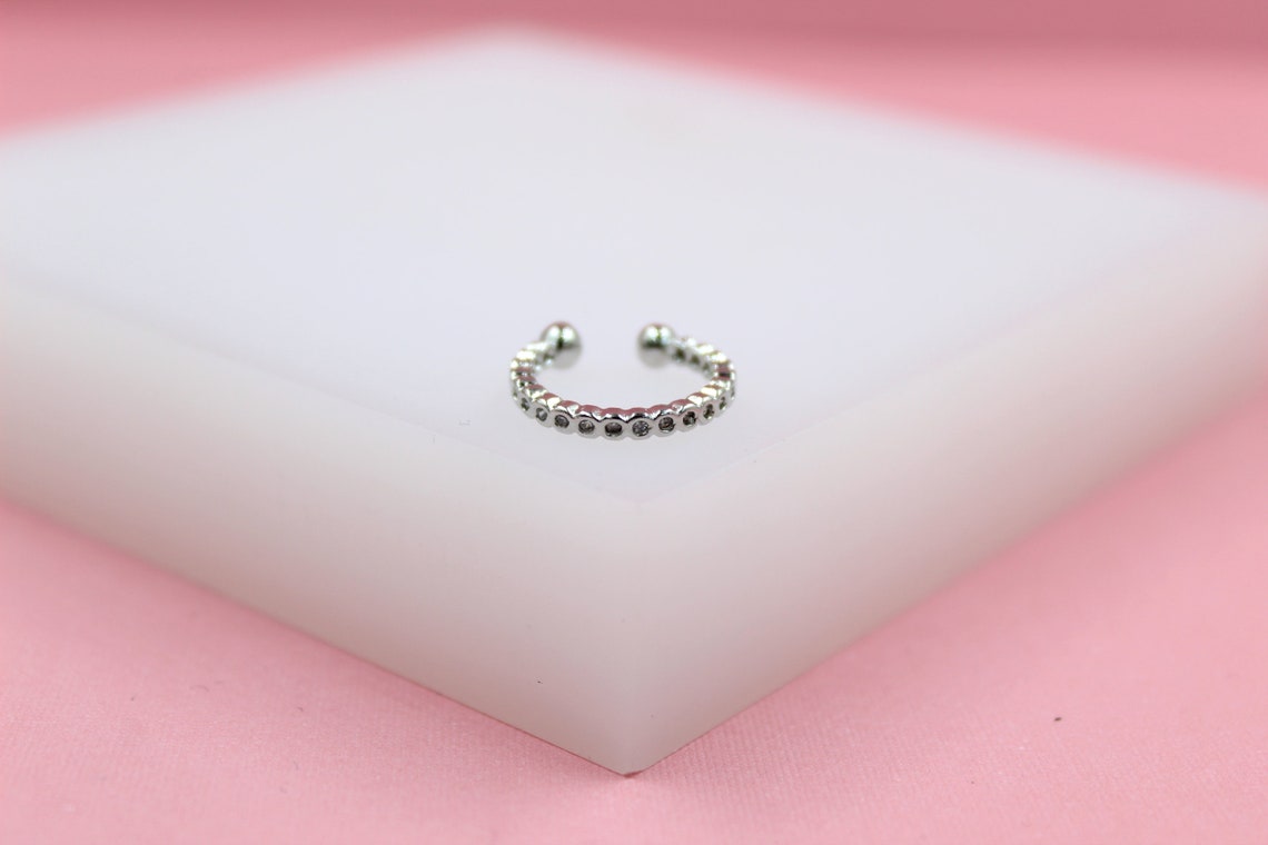 A sparkling silver ear cuff rests on a white display block against a soft pink background.