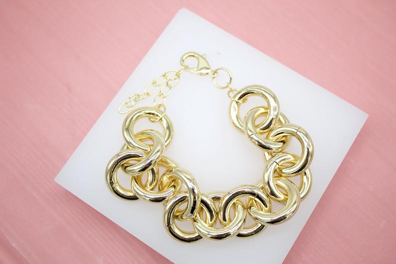 A chunky gold chain link bracelet rests gracefully on a white display card against a soft pink background.