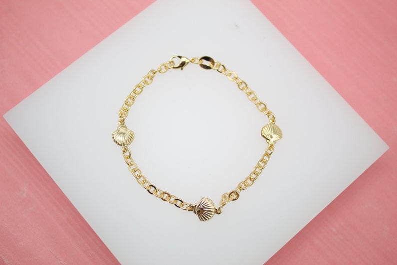 A delicate gold shell charm bracelet rests on a white display surface against a pink background.